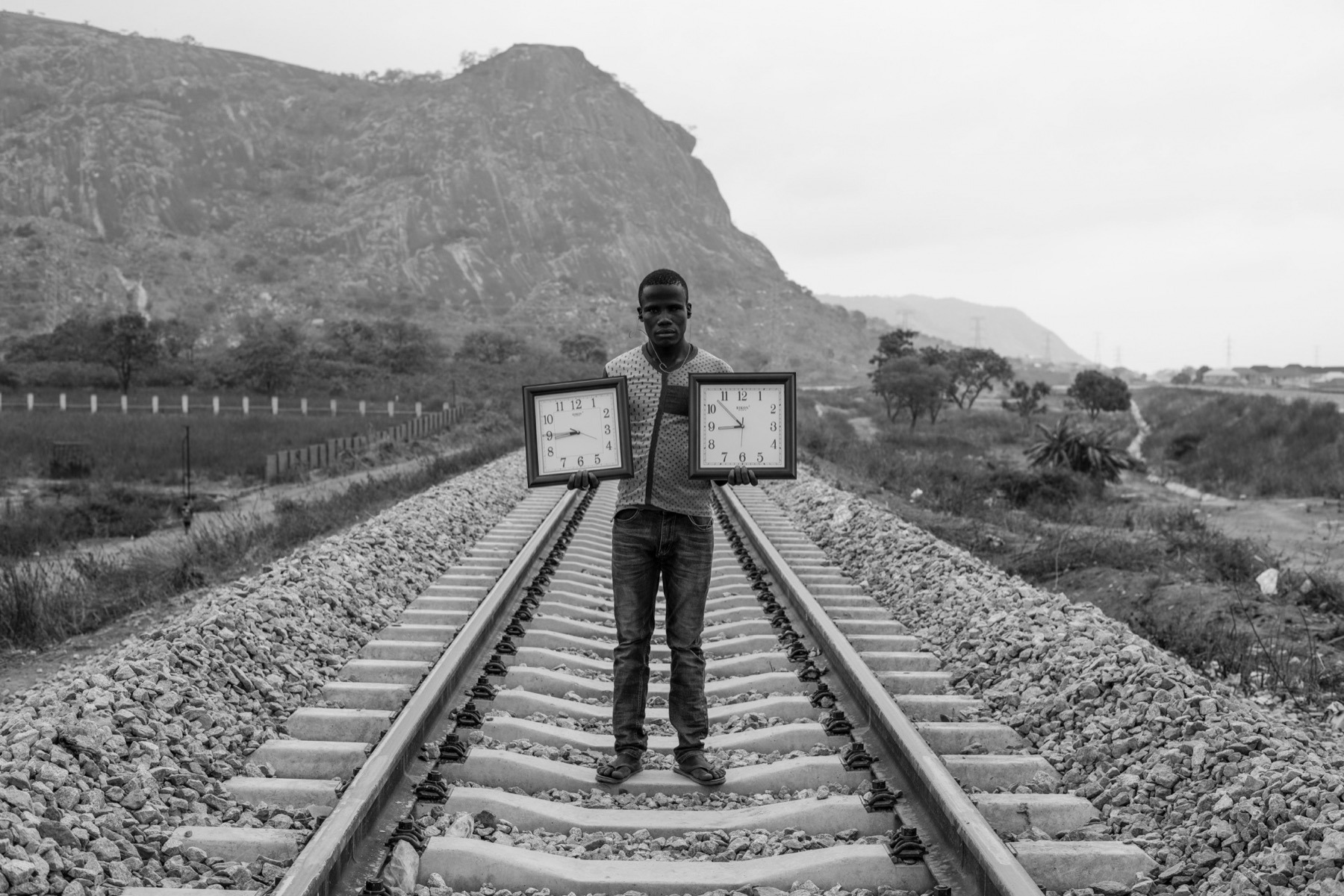 A young man stands on railway tracks holding two framed clocks, a rock outcrop behind him.