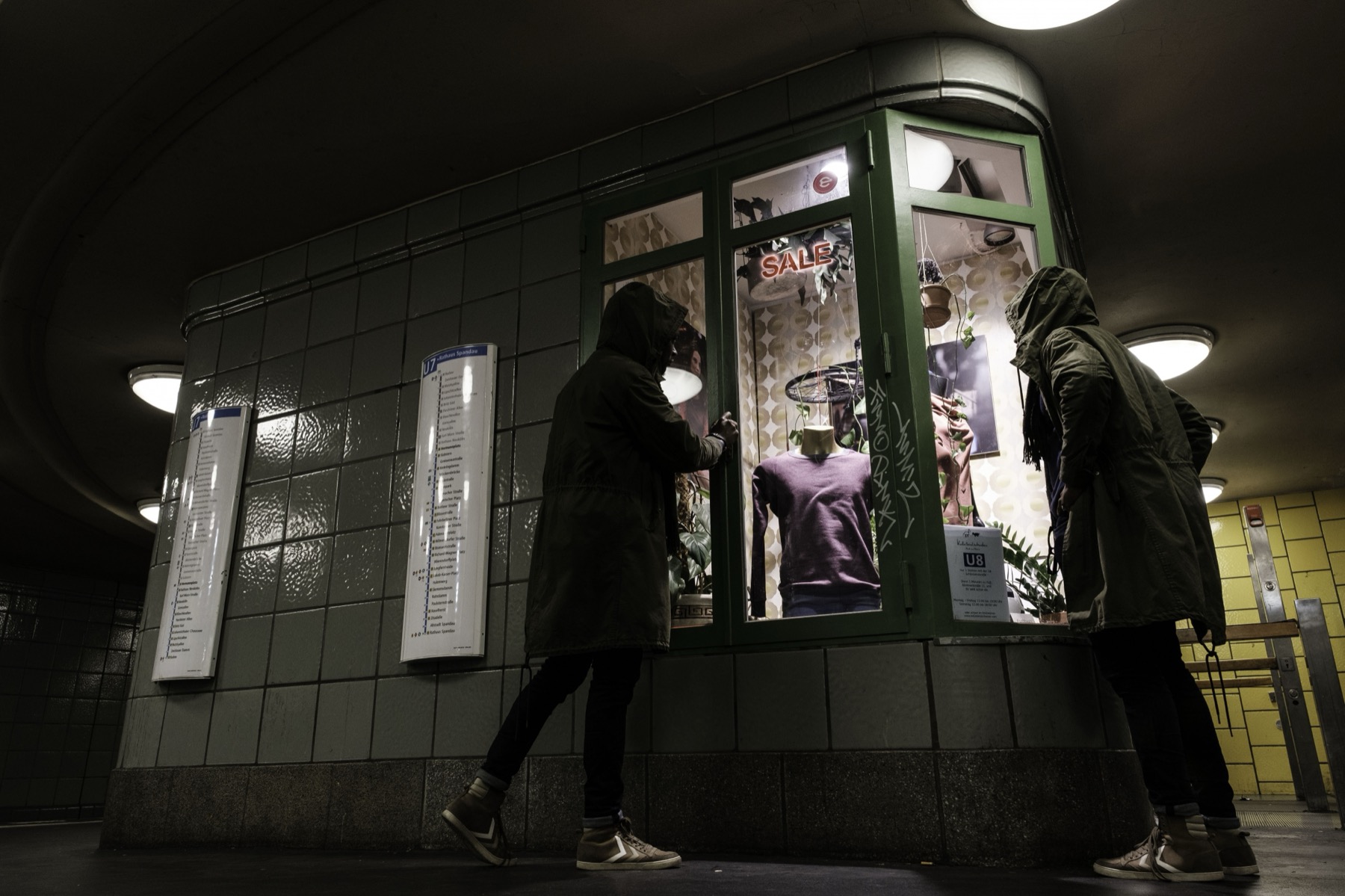 A lit shop window in an underground passage at night. Two figures pass; one turns to look in at the clothes behind the glass.