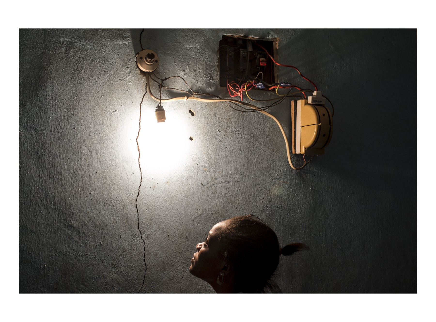 A bare bulb burns on a cracked concrete wall, wired through a tangle of red and yellow cable from an open fuse box to a yellow switch; below and to the right a young woman’s head is in silhouette, turned up towards the light.