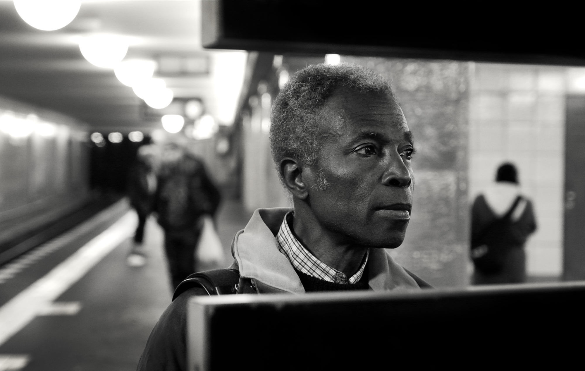 Akinbode Akinbiyi in profile on a Berlin U-Bahn platform, lit by the overhead lamps, figures blurred along the platform behind him.