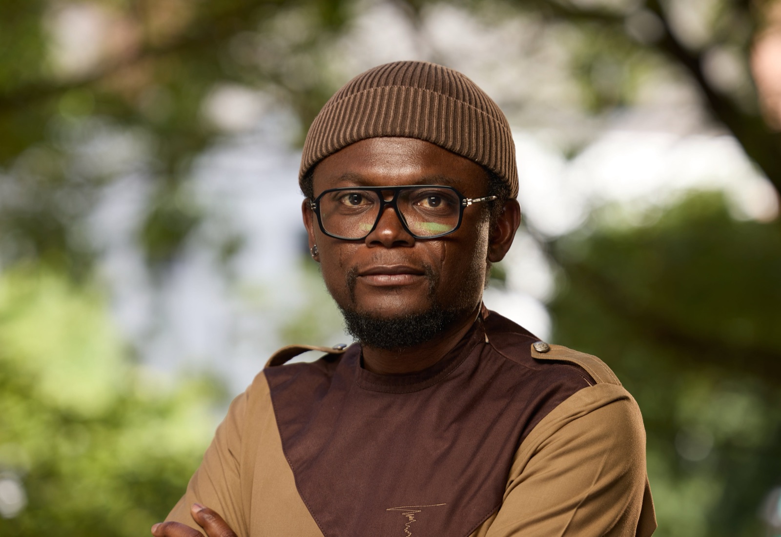 Emeka Okereke outdoors, arms folded, in a brown cap and glasses, looking towards the camera, with sunlit leaves behind him.