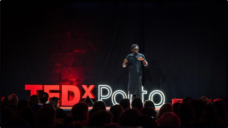 Emeka Okereke speaking on stage before an audience, the lit TEDxPorto sign behind him.